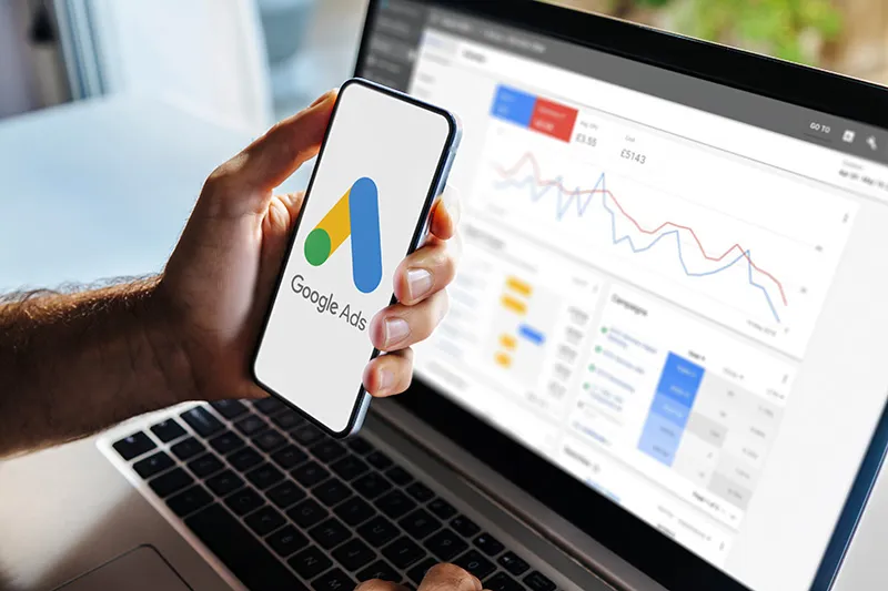 A Google Ads campaign dashboard open on a laptop beside a phone showing the Google Ads logo